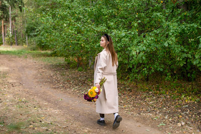 A cute girl in a light coat walks through the autumn forest with a bouquet of flowers