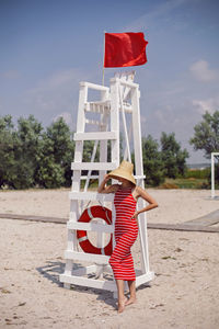 Woman in a red dress stands next to a rescue station with a flag and a lifebuoy on  sea sandy beach