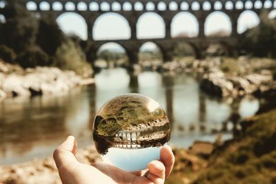 Cropped image of person holding crystal ball with reflection of bridge against sky