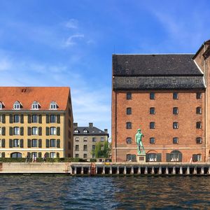 Statue and buildings in front of canal against sky on sunny day