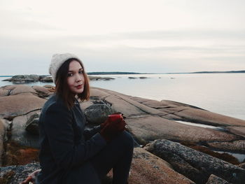 Young woman sitting at sea shore against sky