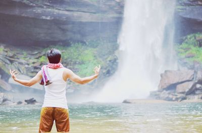 Rear view of man with arms outstretched standing in front of waterfall