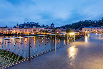 Illuminated bridge over river by buildings against sky at dusk