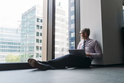 Businessman with laptop sitting on floor by window in office
