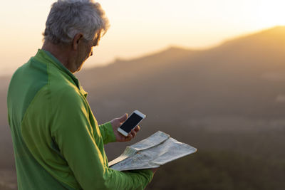 Senior man looking at map and smartphone on top of mountain during sunset