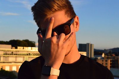 Portrait of young man wearing sunglasses while gesturing horn sign against sky