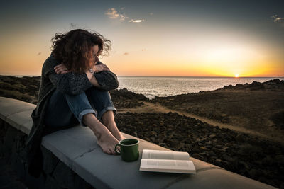 Woman sitting by cup of coffee and book on building terrace against sky