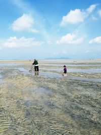 Rear view of grandfather and granddaughter walking at beach against cloudy sky