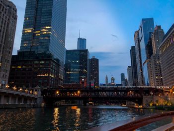 Illuminated bridge over river by buildings against sky at dusk