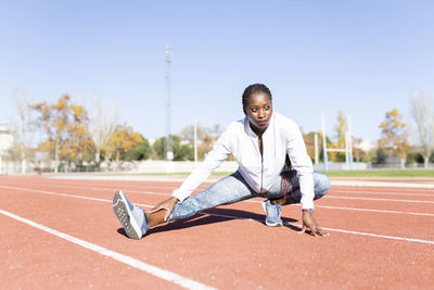 Young sportswoman looking away while doing warm up exercise on sports track against clear sky