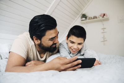 Father and smiling disabled son watching movie over mobile phone while lying on bed at home