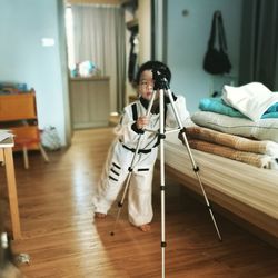 Full length of boy standing by tripod on hardwood floor at home