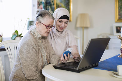 Young female home caregiver with senior woman entering bank using laptop at home