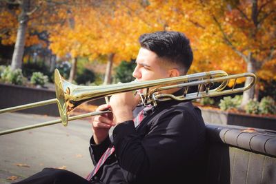 Young man playing trumpet while sitting in park during autumn