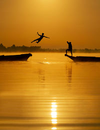 Silhouette men fighting on boats in lake against clear orange sky