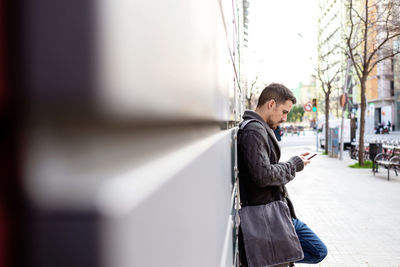 Side view of a bearded man using phone leaning on office building wall