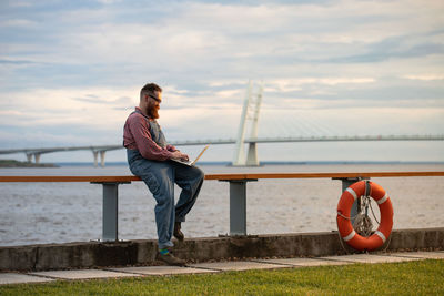 Young man sitting on railing by sea against sky