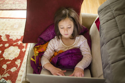 A little girl sits on couch in a pile of blankets working on computer