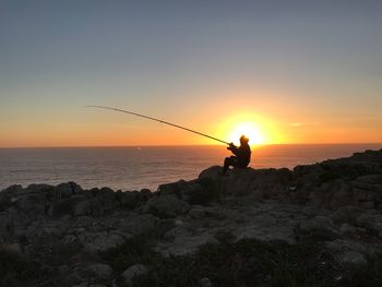 Silhouette fishing rod on rock by sea against sky during sunset