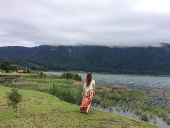 Full length rear view of woman walking on grassy field by lake against cloudy sky