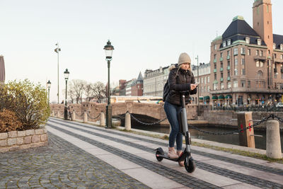 Teenage girl text messaging on smart phone while riding e-scooter on street by canal in city against sky