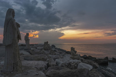 Statue standing on rock by sea against sky during sunrise