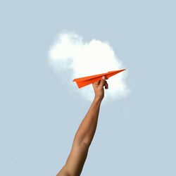 Low angle view of cropped hand holding orange paper rocket against sky