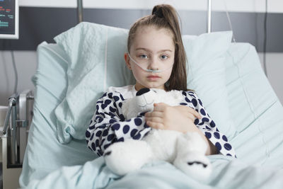 Portrait of girl with oxygen tube embracing teddy bear on bed in hospital