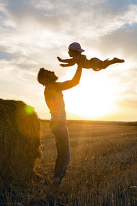 Father throws his son into the sky next to a haystack on a sloping field during sunset