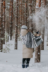 A girl in a striped scarf stands in a winter forest and throws snow in the air.