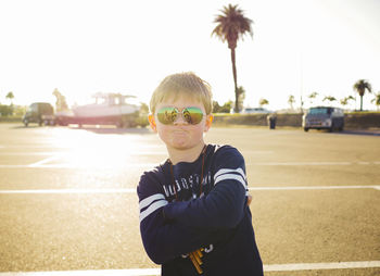 Portrait of confident boy wearing sunglasses while standing arms crossed at parking lot on sunny day