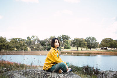 Portrait of woman sitting on rock by lake against sky