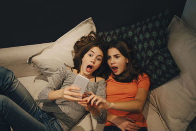 High angle view of friends with open mouth using phone while resting on sofa at home