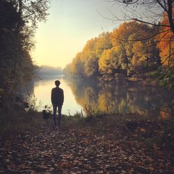 Rear view of man with dog standing at lakeshore by autumn trees