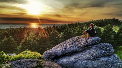 Man sitting on rock against sky during sunset