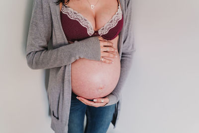 Midsection of pregnant woman in bra standing against wall
