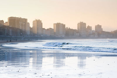 Frozen river by buildings against sky in city during winter