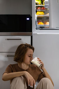 Woman drinking milk while sitting against cabinet at home