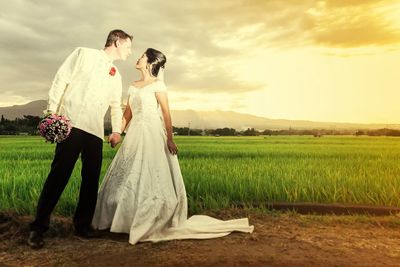 Newlywed couple romancing while standing on field against sky during sunset