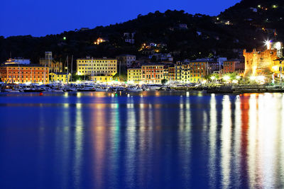 Illuminated buildings in village by sea against silhouette mountain