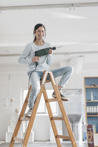 Young woman in her new flat sitting on ladder, holding electric drill