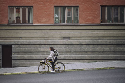 Side view full length of female freelancer moving down on street while commuting through bicycle in city