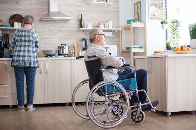 Disabled man sitting on wheelchair with woman in background at home