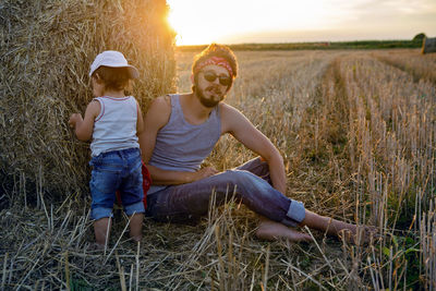 Father and son in t-shirts sitting next to a haystack on a sloping field during sunset