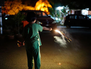 Rear view of girl holding illuminated firework on street in city at night