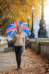 Portrait of woman holding umbrella while walking on footpath during autumn
