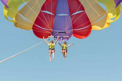 Two female parasailing against bright blue sky in summer with colorful rainbow parachute.