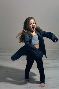 Funny portrait of fashionable girl child in large gray jacket stands against gray wall next  window 