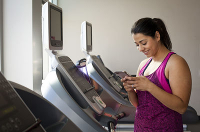 Smiling female using smart phone while standing on treadmill in gym