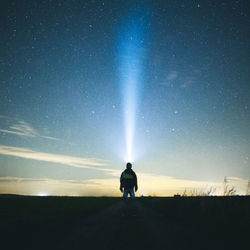 Rear view of silhouette man holding flashlight while standing on field against sky